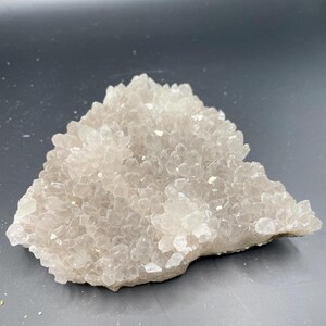May include: A cluster of small, clear quartz crystals in a light beige color. The crystals are densely packed together, forming a textured, irregular shape. The quartz formation sits on a flat, light-colored base.