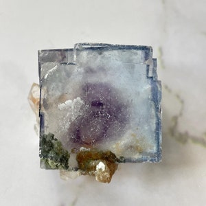 May include: A clear, blue fluorite crystal with purple and green inclusions. The crystal is sitting on a bed of smaller, white crystals.