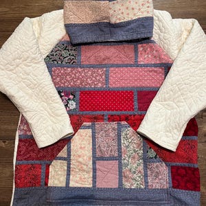 May include: A hooded sweatshirt constructed from a patchwork of various red, pink, and floral patterned fabrics. The garment features long, cream-colored quilted sleeves and a large front kangaroo pocket, all assembled with blue fabric borders.
