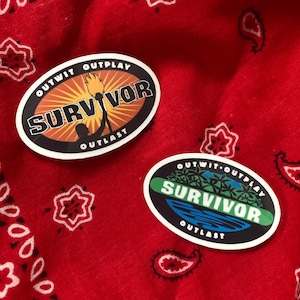 May include: Two oval stickers with the word "SURVIVOR" in bold letters. One sticker features a torch and sunburst design, the other a tropical scene. Both stickers have the phrases "OUTWIT, OUTPLAY, OUTLAST". The stickers are on a red bandana.