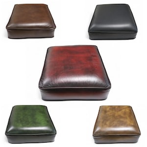 May include: Five square leather ottomans in different colours are shown. The colours are dark brown, black, burgundy, green, and a lighter brown. Each ottoman has a padded top and a stitched border.