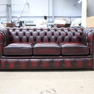 May include: A burgundy leather Chesterfield sofa with a three-cushion design. The sofa features a button-tufted back and arms, rolled armrests, and nailhead trim. The sofa is approximately 213 cm long.
