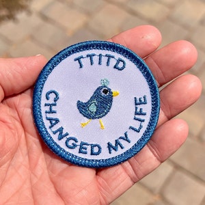 Puede incluir: Un parche bordado azul y blanco con un pequeño pájaro azul con pico y patas amarillas. El texto en el parche dice "TITD Changed My Life".
