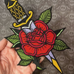 Könnte beinhalten: Eine rote Rose mit einem grünen Stiel und Blättern wird von einem schwarzen und gelben Dolch durchbohrt. Der Dolch hat einen violetten Griff und eine gelbe Klinge.