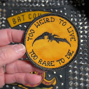 Op de afbeelding: Een gele geborduurde patch met zwarte stiksels. De patch heeft een vleermuis silhouet en de tekst "Too Weird To Live Too Rare To Die".