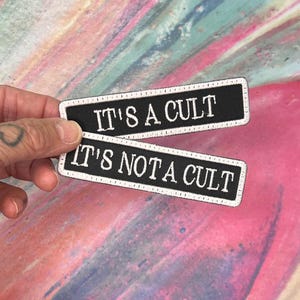 May include: Two rectangular black patches with white text. The top patch reads "IT'S A CULT" and the bottom patch reads "IT'S NOT A CULT". Both patches have a white border.