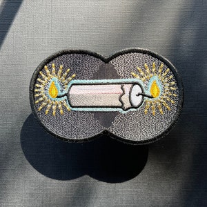 Pode incluir: Um remendo bordado em preto e branco com uma vela e chamas. A vela é branca com um centro rosa e uma borda azul. As chamas são amarelas com um centro branco.