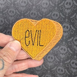 Może przedstawiać: Żółta naszywka w kształcie serca z wyszytym czarnym szwem napisem "EVIL".