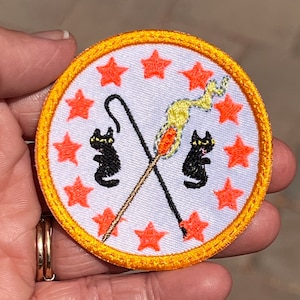Könnte beinhalten: Ein weißer gestickter Aufnäher mit zwei schwarzen Katzen, einem Stock mit einer Flamme und einem Kreis aus orangefarbenen Sternen am Rand. Der Aufnäher hat einen gelben Rand.