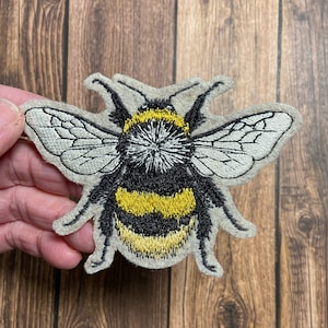May include: A felt embroidered patch of a bumblebee with black and yellow stripes and white wings.