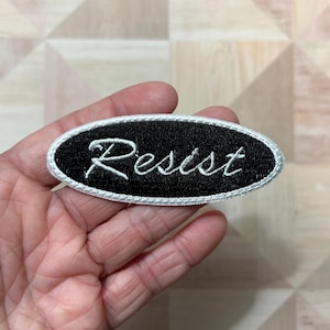 Peut inclure: Écusson ovale noir avec le mot "Resist" brodé en écriture blanche. Le patch a une bordure blanche et est tenu dans une main.