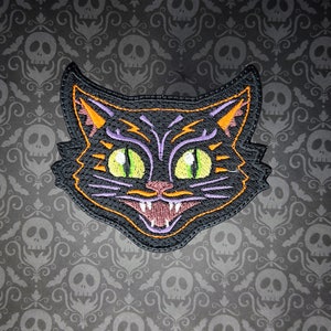 May include: Black embroidered cat face patch with orange and purple details. The cat has green eyes and a wide, toothy grin.