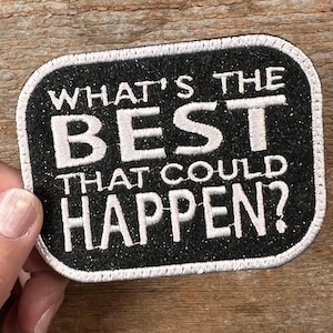 May include: A rectangular black patch with white text reading "WHAT'S THE BEST THAT COULD HAPPEN?". The patch has a glittery finish and a white border, suitable for clothing or accessories.