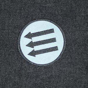 May include: A round patch featuring three black arrows pointing left, set against a light blue background. The patch is bordered by a black stitched edge and is set on a dark gray fabric.