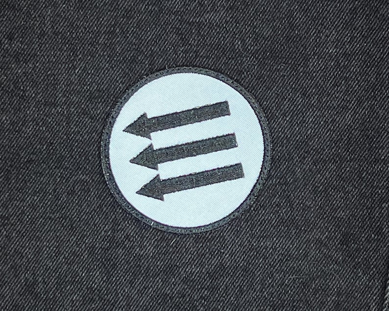 May include: A round patch featuring three black arrows pointing left, set against a light blue background. The patch is bordered by a black stitched edge and is set on a dark gray fabric.