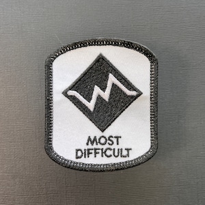 Op de afbeelding: Zwart-witte geborduurde patch met een ruitvorm die een witte bliksem bevat en de tekst "MOST DIFFICULT".
