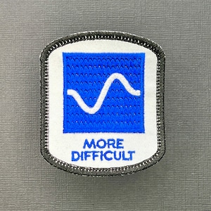Op de afbeelding: Een witte patch met een blauw vierkant met een witte golvende lijn. De tekst "MORE DIFFICULT" staat onder het vierkant.