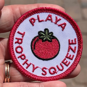 以下が含まれることがあります： トマトと「Playa Trophy Squeeze」という文字が刺繍された赤、白、緑のワッペンです。
