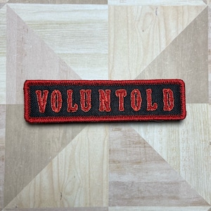 May include: A rectangular black patch with red embroidered text reading "VOLUNTOLD". The patch has a red border and is displayed on a light-coloured, patterned surface. The patch is likely for clothing or accessories.