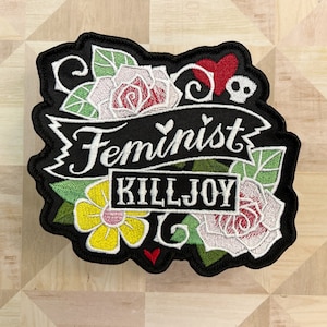 Puede incluir: Parche bordado negro con una pancarta blanca que dice "Feminist Killjoy" rodeado de flores rosas y amarillas y hojas verdes.
