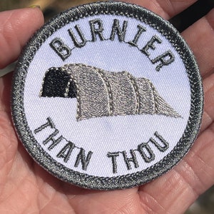 Puede incluir: Un parche bordado blanco con un borde gris. El parche presenta una tienda de campaña con el texto "Burnier Than Thou" alrededor de la tienda.