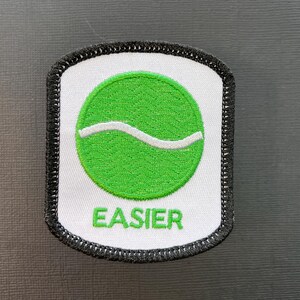 May include: Embroidered patch with a white background and a green circle containing a white wavy line. The word "EASIER" is embroidered in green below the circle. The patch has a black border.
