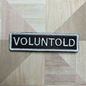 May include: Black and white embroidered patch with the word "VOLUNTOLD" in white lettering.