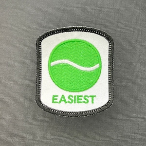 Op de afbeelding: Een witte en groene geborduurde patch met een golfontwerp en het woord "EASIEST" in zwarte tekst.