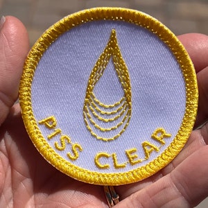 May include: A white round patch with a yellow border and a yellow water drop design. The text "PISS CLEAR" is embroidered in yellow around the water drop.