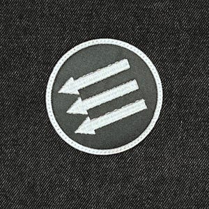 May include: A circular patch with a black background and three white arrows pointing left. The patch has a white border and is set against a dark denim fabric.