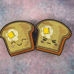 Könnte beinhalten: Zwei gestickte Aufnäher mit Toastscheiben und Butter. Eine Scheibe hat ein lächelndes Gesicht, die andere ein verärgertes Gesicht. Der Toast ist hellbraun mit braunen Rändern, und die Butter ist gelb. Für Kleidung oder Accessoires.