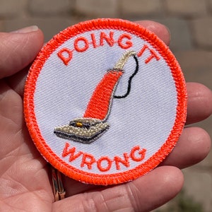 Könnte beinhalten: Orangefarbener und weißer gestickter Bügel-Patch mit dem Text "Doing it Wrong" und einem Cartoon-Bild eines Staubsaugers.