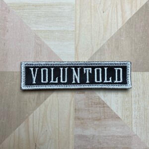 May include: A rectangular black embroidered patch with the word "VOLUNTOLD" in white capital letters. The patch has a white border, suitable for sewing onto garments or accessories.