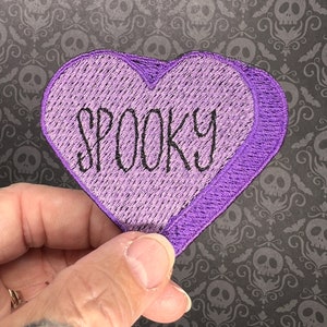 Może przedstawiać: Fioletowa naszywka w kształcie serca z wyszytym czarnym  nicią słowem "SPOOKY".