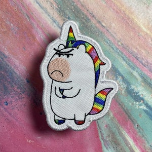 May include: Embroidered patch of a grumpy unicorn with a rainbow mane, tail, and horn. The unicorn is white with a pink snout and is outlined in black. The patch has a white border.