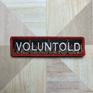 May include: A rectangular black patch with the word "VOLUNTOLD" embroidered in white. The patch has a red border and a flame design in yellow and orange. The patch is on a light brown and white surface.