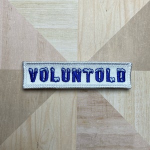 May include: A rectangular patch with the word "VOLUNTOLD" embroidered in blue on a white background. The patch has a silver border and is displayed on a wooden surface.