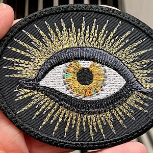 Könnte beinhalten: Gestickter Aufnäher mit einem Augendesign. Das Auge hat eine weiße Iris mit einem gelb-orangen Zentrum, umgeben von einer schwarzen Umrandung und goldenen Strahlen. Der Aufnäher ist kreisförmig mit einem schwarzen Rand.