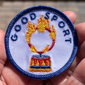 Könnte beinhalten: Ein weißer gestickter Aufnäher mit blauem Rand. Der Aufnäher zeigt ein rotes und gelbes Zirkuszelt mit Flammen, die aus der Spitze ragen. Der Text "GOOD SPORT" ist um den Rand des Aufnähers gestickt.