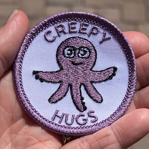 Könnte beinhalten: Lilafarbener gestickter Aufnäher mit weißem Hintergrund. Der Aufnäher zeigt einen Cartoon-Oktopus mit großen Augen und dem Text "Creepy Hugs" um den Oktopus herum.