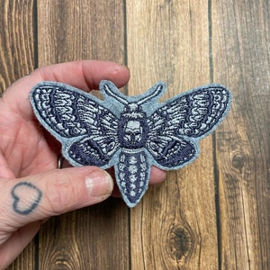 May include: A grey felt embroidered patch of a moth with black stitching. The moth has a skull-like design on its head.