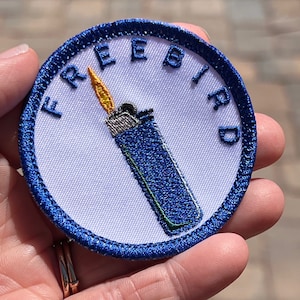 Könnte beinhalten: Ein blauer und weißer gestickter Aufnäher mit dem Wort "FREEBIRD" in einem Kreis um einen blauen Feuerzeug mit einer gelben Flamme.