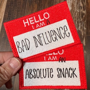 May include: Two red and white name tag patches. The top patch says "Hello I am a Bad Influence". The bottom patch says "Hello I am an Absolute Snack".