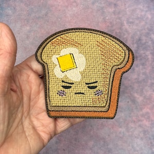 May include: Embroidered patch featuring a slice of toast with a fried egg and a pat of butter. The toast has a grumpy face with stitched details. The patch has a brown border and is likely for clothing or accessories.