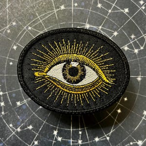 May include: Black oval patch featuring a detailed eye design with gold rays emanating from it. The eye has a white iris and a brown pupil. The patch is set against a background with a constellation pattern.