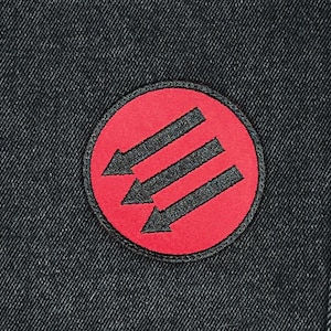 May include: Round patch with a red background and three black arrows pointing down. The patch is sewn onto a dark denim fabric. The patch has a black border.