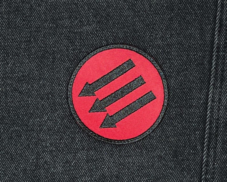 May include: Round patch with a red background and three black arrows pointing down. The patch is sewn onto a dark denim fabric. The patch has a black border.