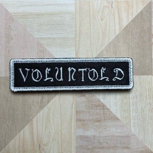May include: A black rectangular patch with the white embroidered word "VOLUNTOLD". The patch has a white border. The patch is set against a light brown and tan patterned surface. The patch is likely for clothing or accessories.