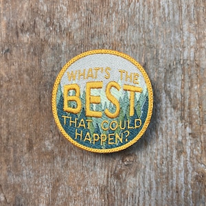 Op de afbeelding: Een ronde, witte geborduurde patch met een gele rand. De patch toont een groene en blauwe berg scène met de tekst "What's the Best That Could Happen?" in geel.