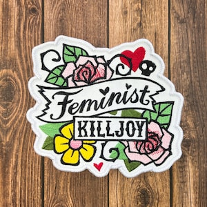 Puede incluir: Parche blanco bordado con una rosa rosa y roja, hojas verdes, una flor amarilla y una pancarta que dice "Feminist Killjoy".